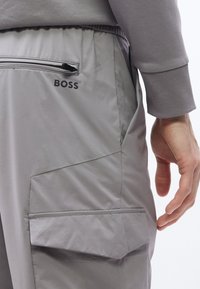 Κοντινό πλάνο γκρι παντελονιών με τσέπη με φερμουάρ και τσέπη με καπάκι, που δείχνει ελαστική μέση και χέρι που ακουμπάει δί πλα στο ισχίο.