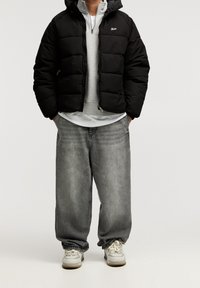 Chaqueta acolchada negra con capucha, sudadera gris debajo, vaqueros grises sueltos y zapatillas blancas con detalles claros. Cierre delantero con cremallera, textura acolchada.