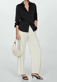 Une chemise noire à boutons, un pantalon large crème, des sandales à talons noires et un sac à main courb é en blanc cassé. La chemise a des manches retroussées.