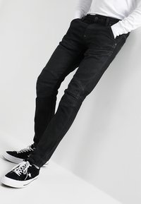 Mustat farkut, joissa on slim fit -leikkaus, polvien ompeluyksityiskohdat ja hieno haalistuma. Yhdistetty mustien tennareiden ja valkoisen pitkähihaisen paidan kanssa.