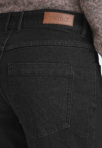 Jeans de mezclilla negra con un parche de cuero etiquetado como "oxmo", con un bolsillo trasero y detalles de costura visibles. Textura suave y diseño ajustado.