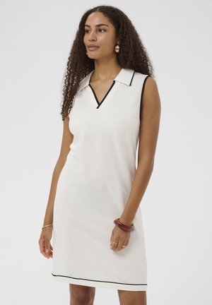 Kvinde med krøllet hår iført hvid ærmeløs kjole med sort kant, guldringeøreringe og vinrødt armbånd, stående mod en ensfarvet baggrund.