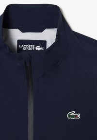 Marineblauwe Lacoste sportjack met een hoge kraag, ritssluiting en een groen krokodillenlogo op de borst; gladde textuur en atletisch ontwerp.