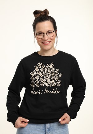 Ung kvinde med briller og håret sat op i en knold iført sort sweatshirt med beige Henri Matisse bladdesign og lyseblå jeans, smilende.