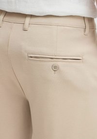 Les pantalons beiges ajustés présentent une texture lisse, une coupe droite et une poche arrière à bouton unique. La taille est équipée de passants de ceinture.