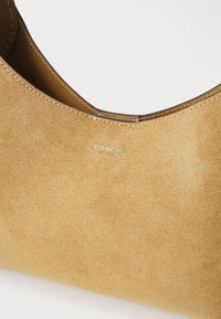 Sac à main en daim tan avec une forme arrondie, présentant un intérieur lisse et des coutures contrastées. Logo embossé près du bord supérieur.