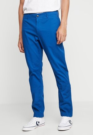 Mężczyzna w jasnoniebieskich obcisłych spodniach, białych trampkach z granatowymi logo gwiazdek oraz białej koszuli włożonej do środka, stojący na jednolitym tle.