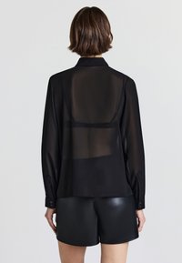 Blusa nera trasparente con maniche lunghe, colletto a punta e polsini con bottoni. Indossata sopra pantaloni corti in finta pelle nera. Texture liscia, leggermente traslucida.