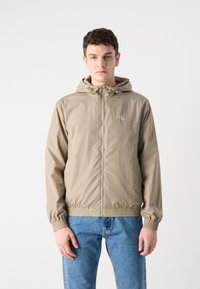 Lys beige hættetrøje med elastiske manchetter og kant, med en for-lynget lynlås og et lille logo på brystet. Sammen med blå jeans.