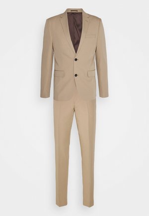 Costume beige avec une veste à revers, deux boutons et une poche, associée à un pantalon slim en tissu et couleur assortis.