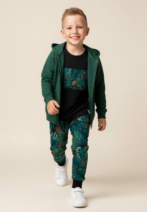 Uśmiechnięty chłopiec w ciemnozielonej bluzie na zamek, czarnej koszulce z tropikalnym nadrukiem, spodniach z tym samym tropikalnym wzorem i białych trampkach.