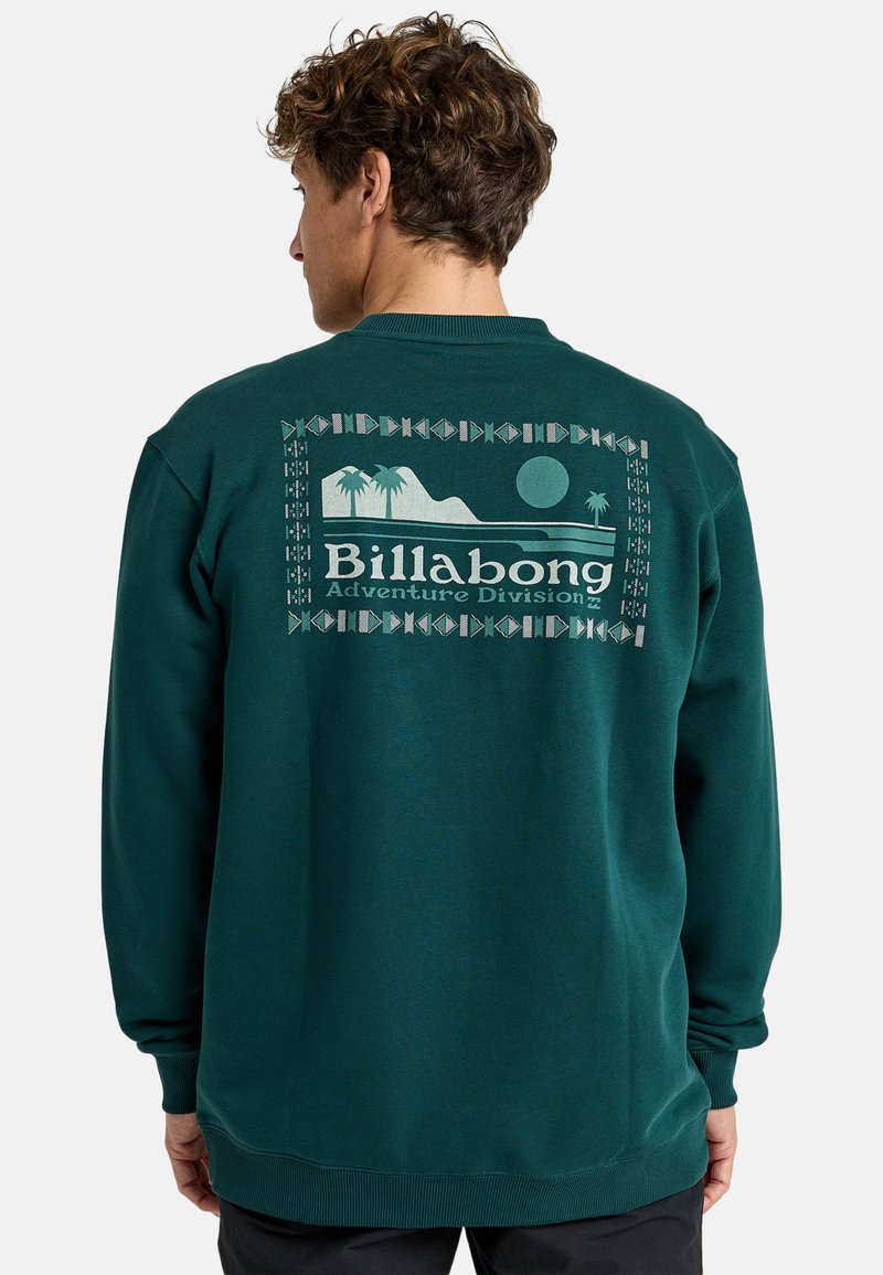Zielona bluza z polaru z okrągłym dekoltem. Posiada dużą, jasnoniebieską grafikę na plecach, przedstawiającą góry, palmy i tekst.