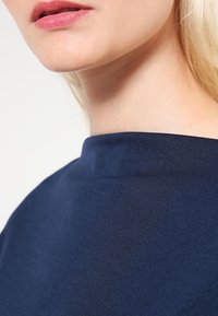 Námořnická modrá halenka s hladkým, elastickým materiálem, s vysokým kulatým výstřihem a minimalistickým designem, který odhaluje část krku a obličeje.