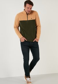 Pulover od flisa s četvrtastim zatvaračem, sa bež gornjim dijelom i maslinasto zelenim donjim dijelom. Mali izvezeni logo na grudima, mekana tekstura.