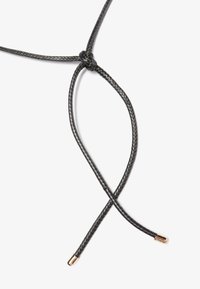 Cordon tressé noir avec un nœud, doté de pointes en métal doré. Le cordon est flexible et lisse au toucher, formant une forme de X au centre.