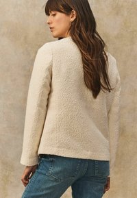 Cremefarbene strukturierte Jacke mit langen Ärmeln. Verfügt über einen geraden Saum und eine lockere Passform, kombiniert mit blauen Jeans.