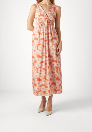 Femme portant une robe maxi sans manches à col en V avec un motif floral orange, rose et blanc et des talons pointus beige sur fond blanc.