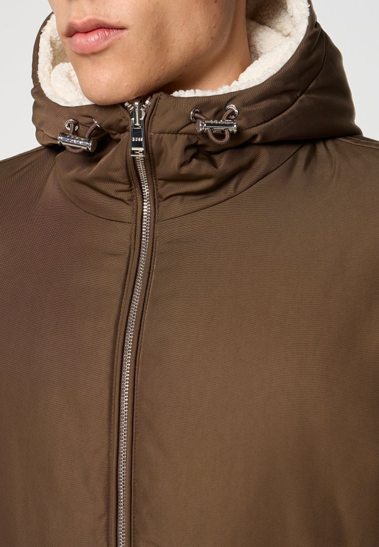 Veste zippée marron avec une coque extérieure texturée et une doublure en polaire crème douce. Comprend une capuche avec cordon de serrage et des accents métalliques.