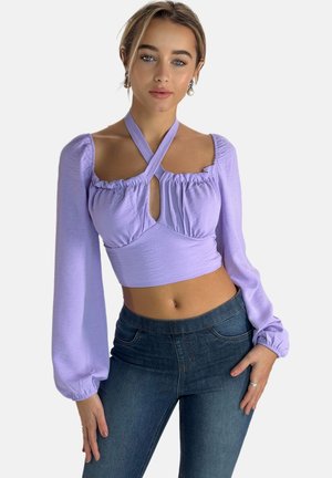 Liliowa crop top z bufkami, z detalem w kształcie dziurki, w fasonie na krzyż. Zestawiona z ciemnoniebieskimi dopasowanymi dżinsami.