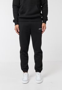 Osoba v černých sportovních kalhotách s bílým logem na stehně, černé mikině s kapucí a bílých teniskách, stojící na bílém pozadí.