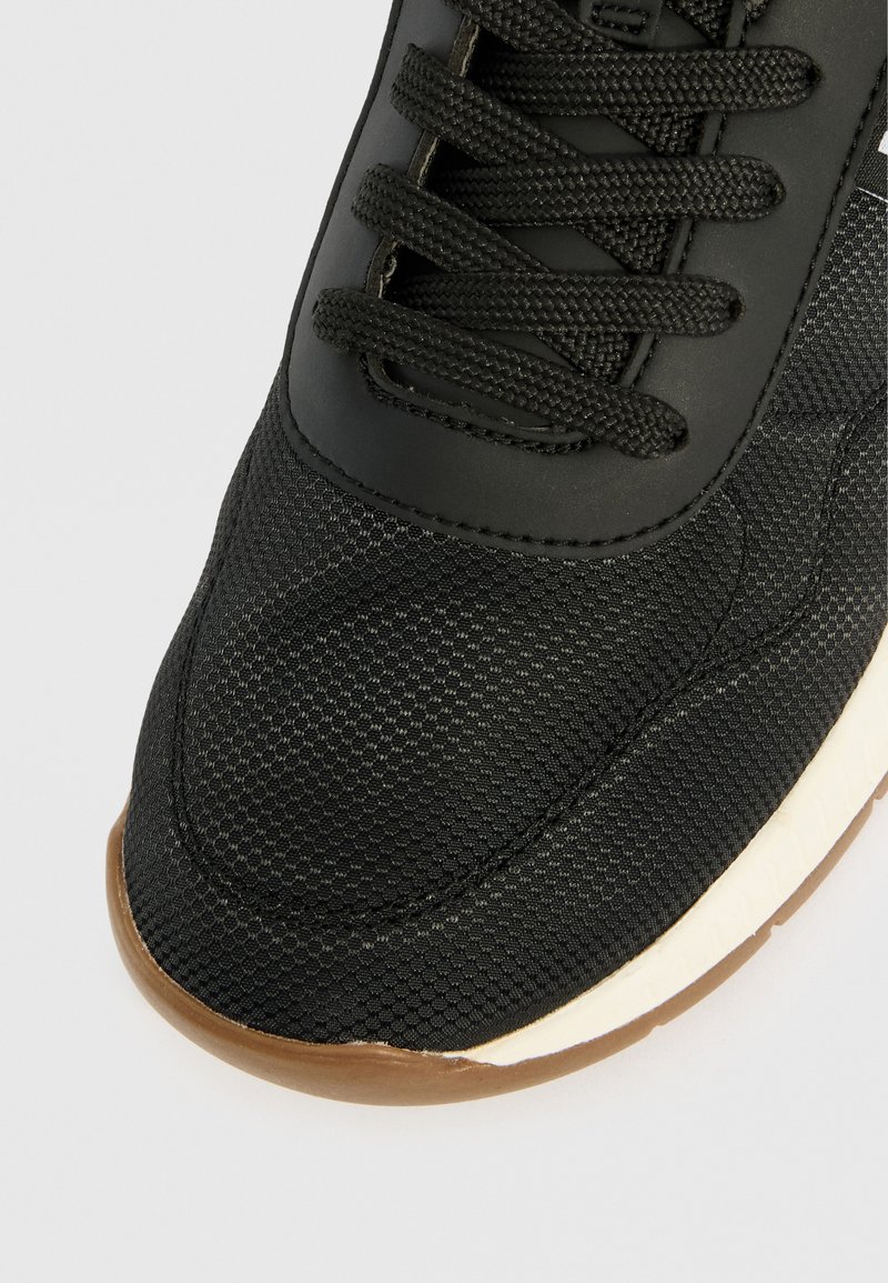 Gros plan sur l'orteil d'une sneaker noire avec un tissu texturé, des lacets noirs, une semelle intermédiaire blanche et une semelle extérieure en caoutchouc marron.