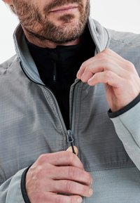 Grå zip-jakke med en tekstureret overflade og kontrasterende sort inderside, der har en delvis lynlås og et close-up af hænder, der justerer den.