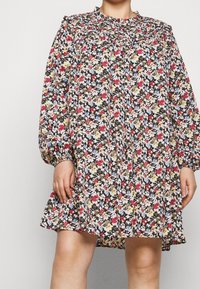 Kukallinen mekko mustalla taustalla, jossa on pieniä punaisia, vaaleanpunaisia, keltaisia ja sinisiä kukkia. Pitkät hihat, rypistetty kaula-aukko ja levenevä helma.