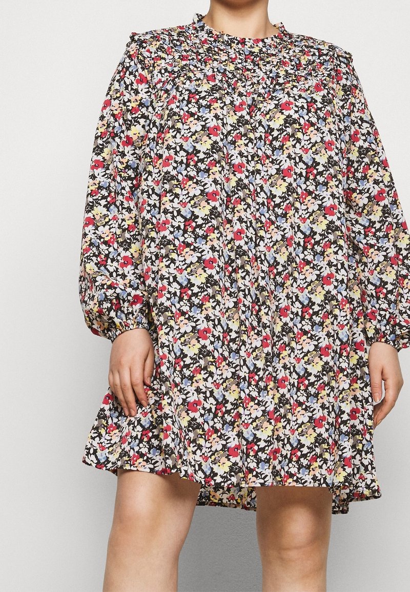 Kukallinen mekko mustalla taustalla, jossa on pieniä punaisia, vaaleanpunaisia, keltaisia ja sinisiä kukkia. Pitkät hihat, rypistetty kaula-aukko ja levenevä helma.