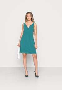 Vestido verde-água com decote em V e detalhe de rendas, corpete ajustado e saia fluida. Usado com saltos verdes e uma carteira branca.