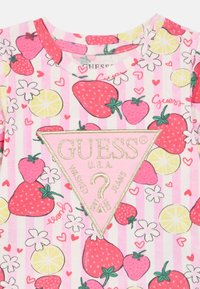 Ροζ πουκάμισο με κοντά μανίκια και σχέδιο με φράουλες και λεμόνια, με ένα τρίγωνο λογότυπο με χρυσή περίγραμμα που γράφει "GUESS U.S.A." και ένα ερωτηματικό.