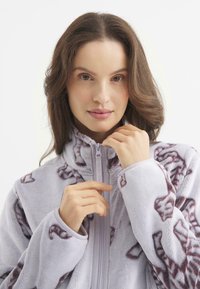 Polarowa kurtka w jasnoszarym kolorze z bordowym abstrakcyjnym wzorem. Posiada wysoki kołnierz, pełny zamek z przodu oraz miękką fakturę.