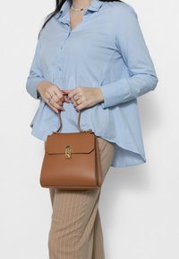 Borsa a mano in pelle marrone con un design strutturato, chiusura in metallo dorato e manici corti, abbinata a una camicia azzurra e pantaloni a righe.