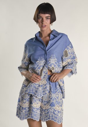 Donna che indossa un completo di camicia e pantaloncini blu con ricamo floreale in pizzo beige, in piedi con le mani vicino alla vita, sfondo neutro.