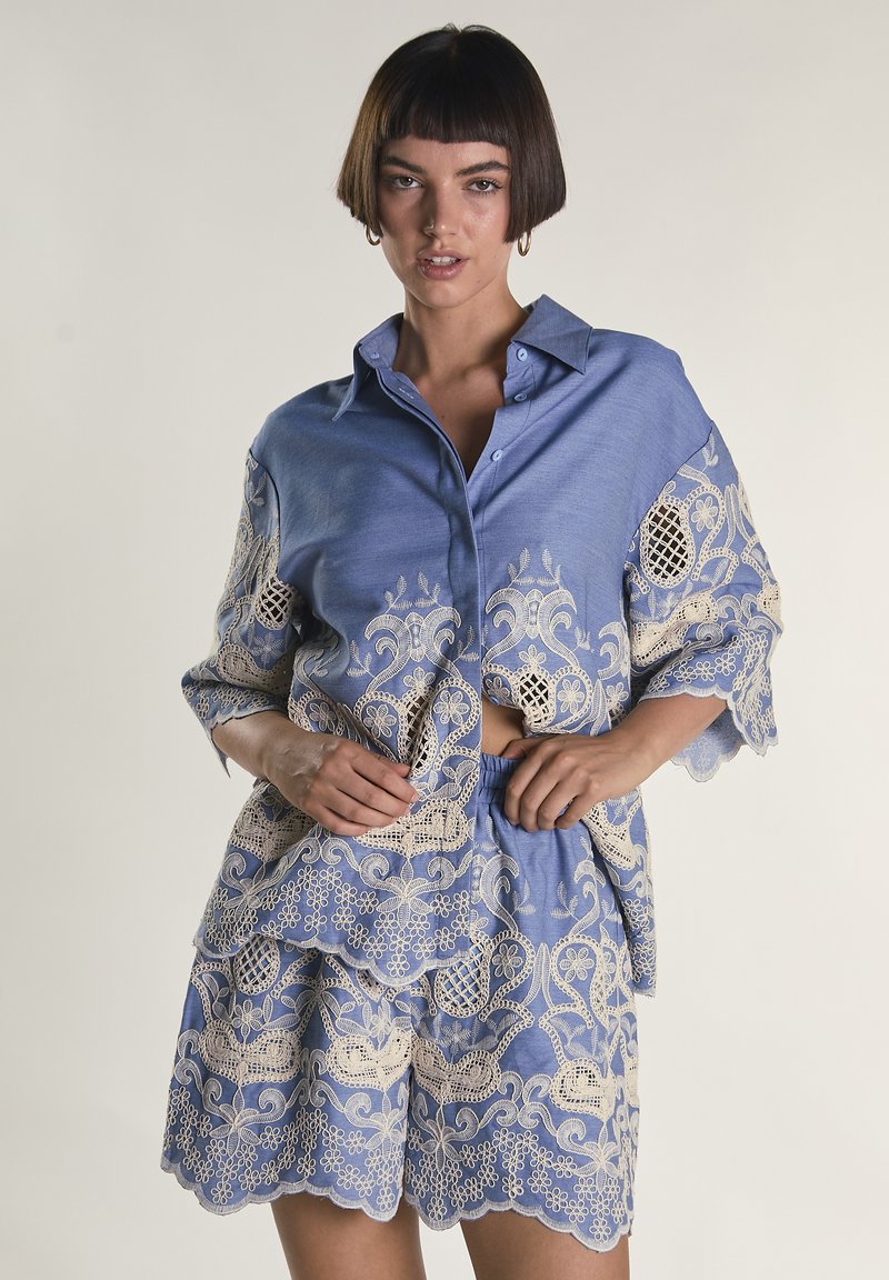 Kvinde iført blåt sæt med skjorte og shorts med beige blomsterbroderi i blonder, stående med hænderne nær taljen, neutral baggrund.