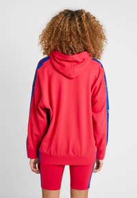 Röd hoodie med blå sidrand, utrustad med huva och löst sittande passform. Baksidan har en slät yta utan framträdande dekorationer.