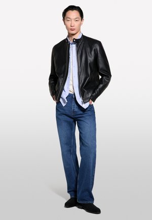 Junger Mann steht mit den Händen in den Taschen, trägt eine schwarze Lederjacke, hellblaues Hemd, blaue Jeans und schwarze Schuhe vor einfachem Hintergrund.
