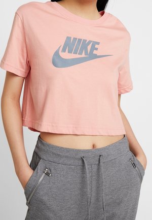 Osoba ubrana w różowy krótki t-shirt Nike oraz szare spodnie dresowe z kieszeniami zapinanymi na zamek, ręce w kieszeniach, widoczny tors.