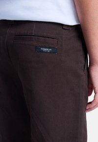 Pantalones marrones de tela texturizada, con un bolsillo trasero que lleva una etiqueta negra que dice "ELEMENT CO. SINCE 1992." Detalles de costura sutiles.