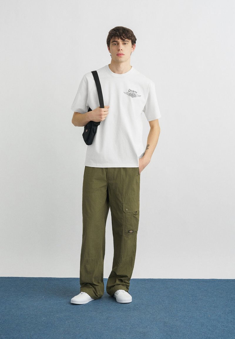 Jeune homme portant un T-shirt blanc à motif, un pantalon cargo vert olive, des baskets blanches et un sac bandoulière noir, debout sur un tapis bleu.