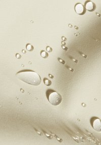 Beige Stoff mit einer glatten Textur, auf dem verstreut klare Wassertropfen unterschiedlicher Größe liegen, die das Licht reflektieren.