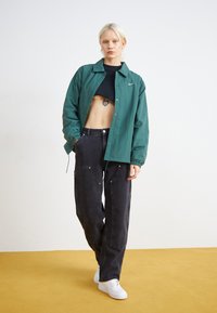 Zöld, könnyű dzseki gombos elülső résszel és rugalmas mandzsettákkal, fekete, bő szárú nadrággal, arany díszítéssel és fehér sportcipőkkel párosítva.