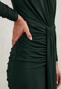 Tume roheline voltitud kleit, mis on valmistatud pehmest kangast, pikkade varrukatega, seotud vöökoht ja küljetasku. Sile tekstuur koos voolavate aktsentidega.