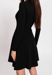 Czarna sukienka z długim rękawem, dopasowanym gorsetem i rozkloszowanym dołem. Gładka faktura z lekkim połyskiem. Proste, czyste linie bez widocznych wzorów.