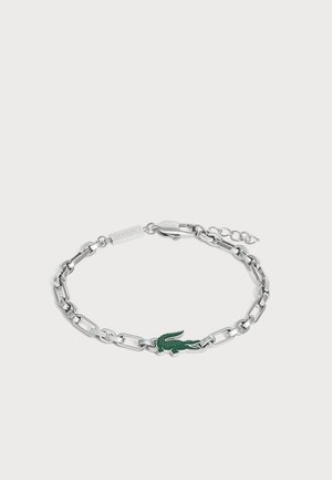 Bracelet chaîne en argent avec un pendentif logo crocodile vert et une fermeture à mousqueton sur fond gris clair.