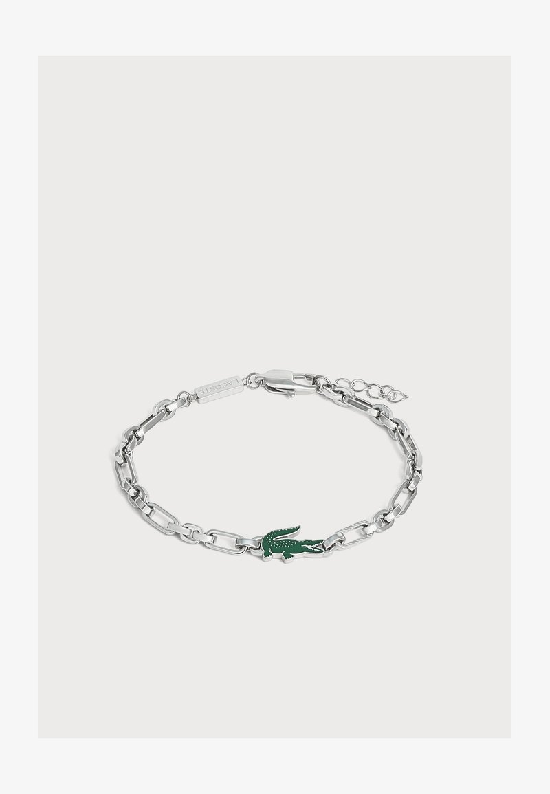 Bracelet chaîne en argent avec un pendentif logo crocodile vert et une fermeture à mousqueton sur fond gris clair.