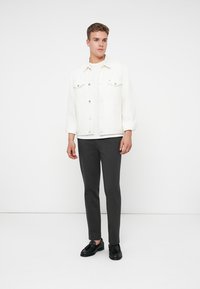 Fiatal férfi fehér farmerdzsekit, fehér pólót, sötétszürke slim fit nadrágot és fekete rojtossal díszített loafert visel, miközben egy fehér háttér előtt áll.