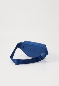 Fanny pack de tecido azul com fecho de zipper, alça ajustável e textura suave. Design compacto com forma arredondada.