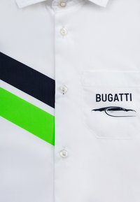 Camicia bianca con bottoni a polo, caratterizzata da righe diagonali verdi e nere, una tasca sul petto con un logo e bottoni di forma perlacea. Realizzata in tessuto liscio.