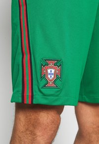 Pantalones cortos deportivos verdes con un escudo bordado de Portugal, con franjas negras y rojas a los lados. Textura suave y ajuste relajado.