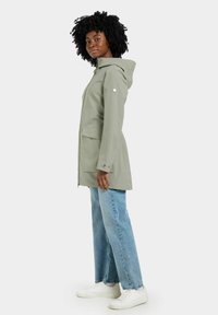 Veste imperméable vert clair avec capuche, fermeture éclair à l'avant et deux poches latérales, associée à un jeans coupe décontractée et des baskets blanches.